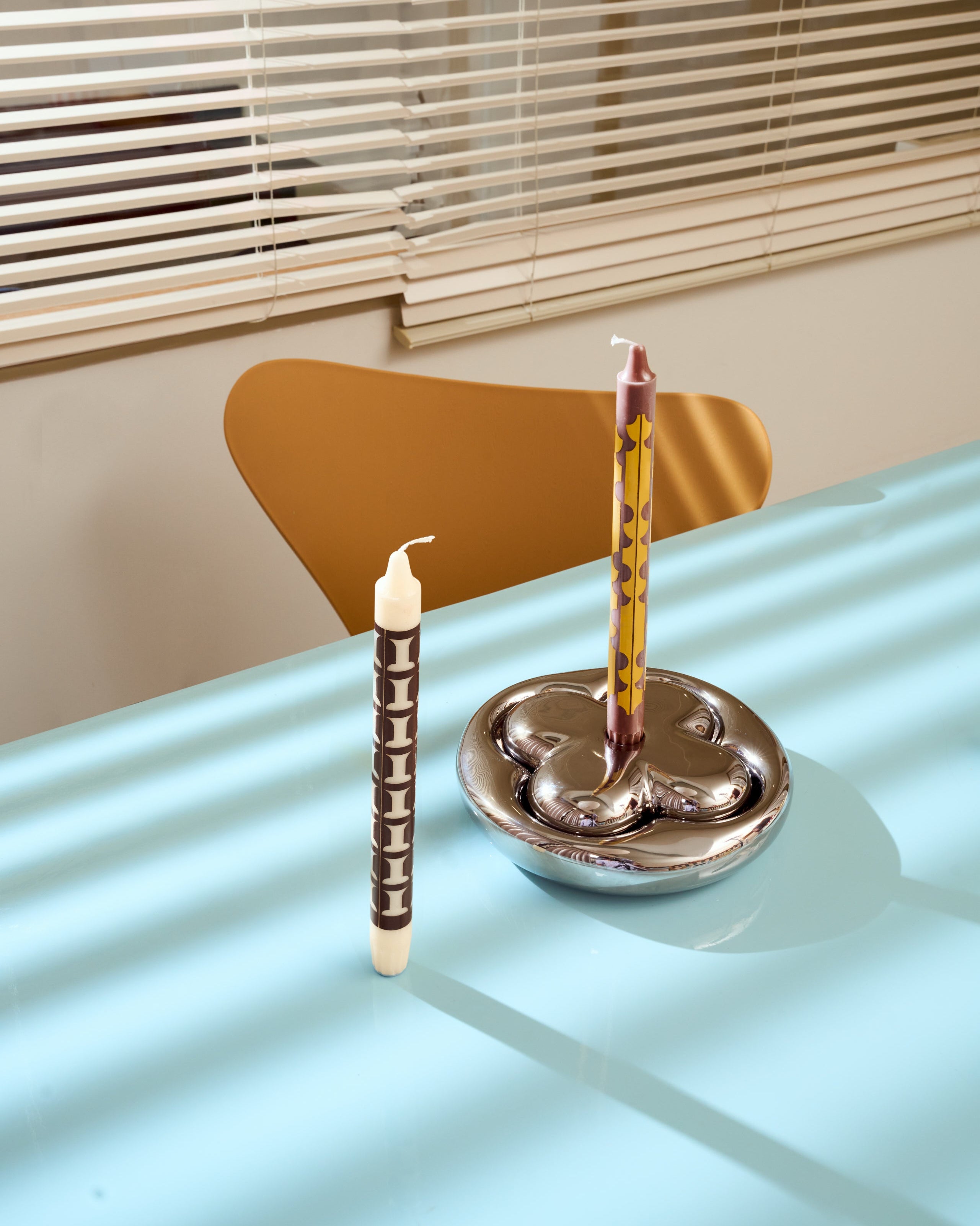 Two ALEO Graphic candles on a glass table with a metallic Folia holder, blinds in the background