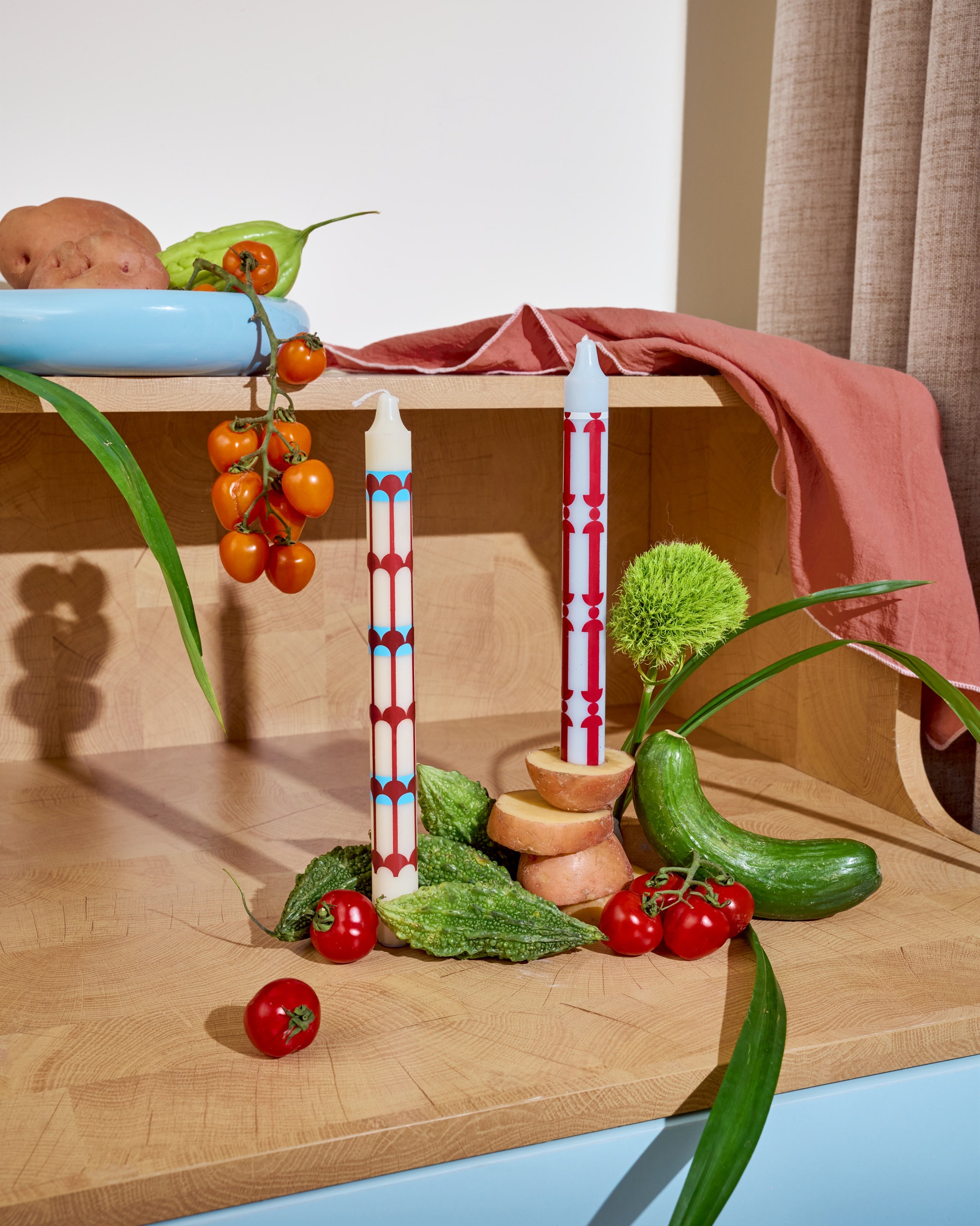 Two candles with decorative patterns on a wooden surface with fruits and vegetables.
